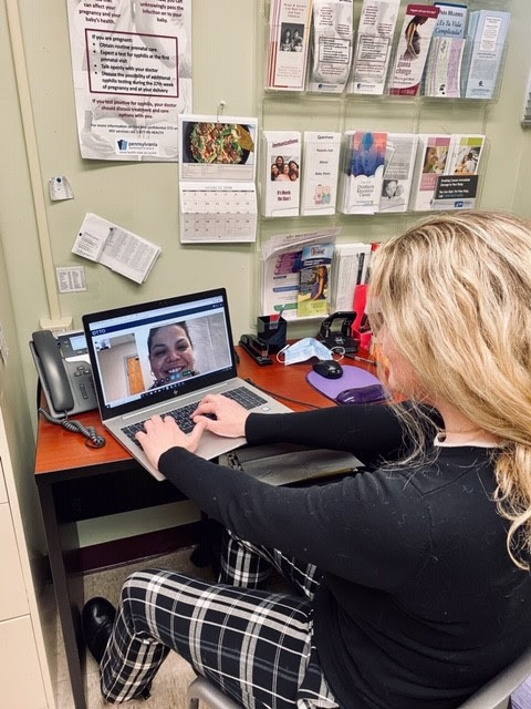 A female provider sits at a cluttered desk with pamphlets on a bulletin board above her. The provider is conducting a telehealth visit on her laptop with a smiling patient.